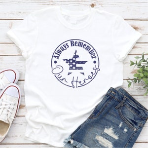 May include: White t-shirt with a blue distressed circle graphic that says "Always Remember Our Heroes" with a star in the center that has an American flag design.