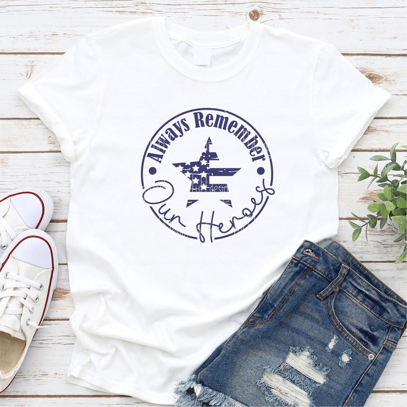 May include: White t-shirt with a blue distressed circle graphic that says "Always Remember Our Heroes" with a star in the center that has an American flag design.