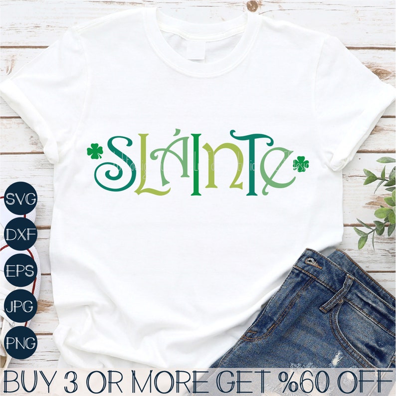 Puede incluir: Camiseta blanca con la palabra "Sl&aacute;inte" en letras verdes, amarillas y azules con acentos de tr&eacute;bol. El texto est&aacute; en una fuente decorativa. La camiseta est&aacute; sobre un fondo de madera con un par de pantalones vaqueros azules.