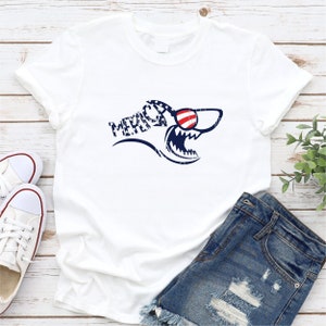 May include: White t-shirt with a blue shark graphic wearing red, white, and blue sunglasses. The shark graphic says "MERICA" in blue letters.