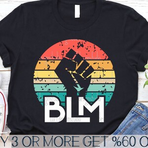Puede incluir: Camiseta negra con un gráfico retro de un puño levantado frente a un fondo de colores arcoíris. El texto "BLM" está debajo del gráfico.