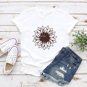 May include: A white t-shirt with a brown sunflower graphic design. The t-shirt is laid out on a wooden surface with a pair of white sneakers and denim shorts.