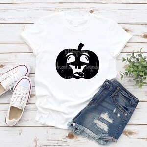 Op de afbeelding: Een wit T-shirt met een zwarte afbeelding van een pompoen met een grappig gezicht. De pompoen heeft zijn ogen opgerold en zijn tong uitsteken. Het T-shirt ligt op een houten oppervlak met een paar witte sneakers en een spijkerbroek.