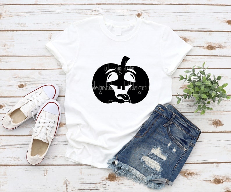 Op de afbeelding: Een wit T-shirt met een zwarte afbeelding van een pompoen met een grappig gezicht. De pompoen heeft zijn ogen opgerold en zijn tong uitsteken. Het T-shirt ligt op een houten oppervlak met een paar witte sneakers en een spijkerbroek.