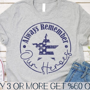 May include: A gray t-shirt with a blue circle design featuring a star with an American flag pattern. The text "Always Remember Our Heroes" is written around the star. The text "BUY 3 OR MORE GET %60 OFF" is written at the bottom of the image.