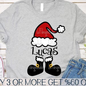 May include: A gray t-shirt with a red and white Santa hat and the name "Lucas" in black text. The Santa hat has a white pom-pom and the name is in a playful font. The t-shirt also features black boot-shaped designs with white trim.