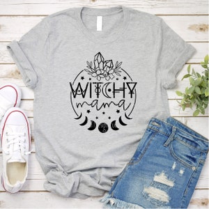 May include: Gray t-shirt with a black graphic design of a circle with the text "Witchy mama" inside. The circle is surrounded by stars, moons, and crystals.