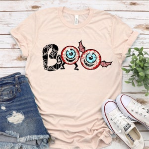 Może przedstawiać: Jasnor&oacute;żowy t-shirt z czarno-białym nadrukiem słowa "BOO" z dwoma kresk&oacute;wkowymi oczami. Oczy mają czerwone powieki, niebieskie tęcz&oacute;wki i białe źrenice. "O" w BOO jest wykonane z jednego oka z wychodzącym z boku skrzydłem nietoperza. Drugie oko służy jako drugie "O".