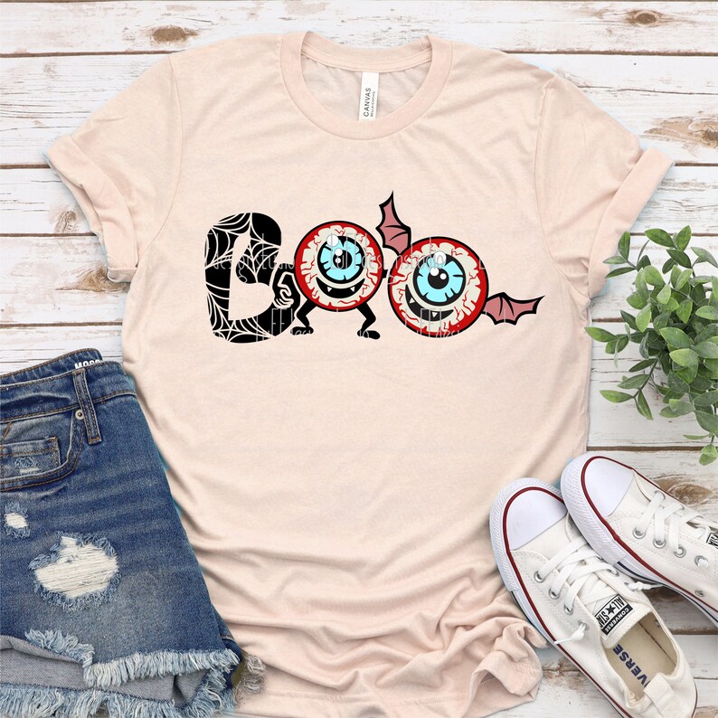 Może przedstawiać: Jasnor&oacute;żowy t-shirt z czarno-białym nadrukiem słowa "BOO" z dwoma kresk&oacute;wkowymi oczami. Oczy mają czerwone powieki, niebieskie tęcz&oacute;wki i białe źrenice. "O" w BOO jest wykonane z jednego oka z wychodzącym z boku skrzydłem nietoperza. Drugie oko służy jako drugie "O".