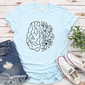 May include: A light blue t-shirt with a black and white graphic of a brain with flowers on one side. The shirt is laid flat on a wooden surface with a pair of white sneakers and a pair of blue denim shorts.