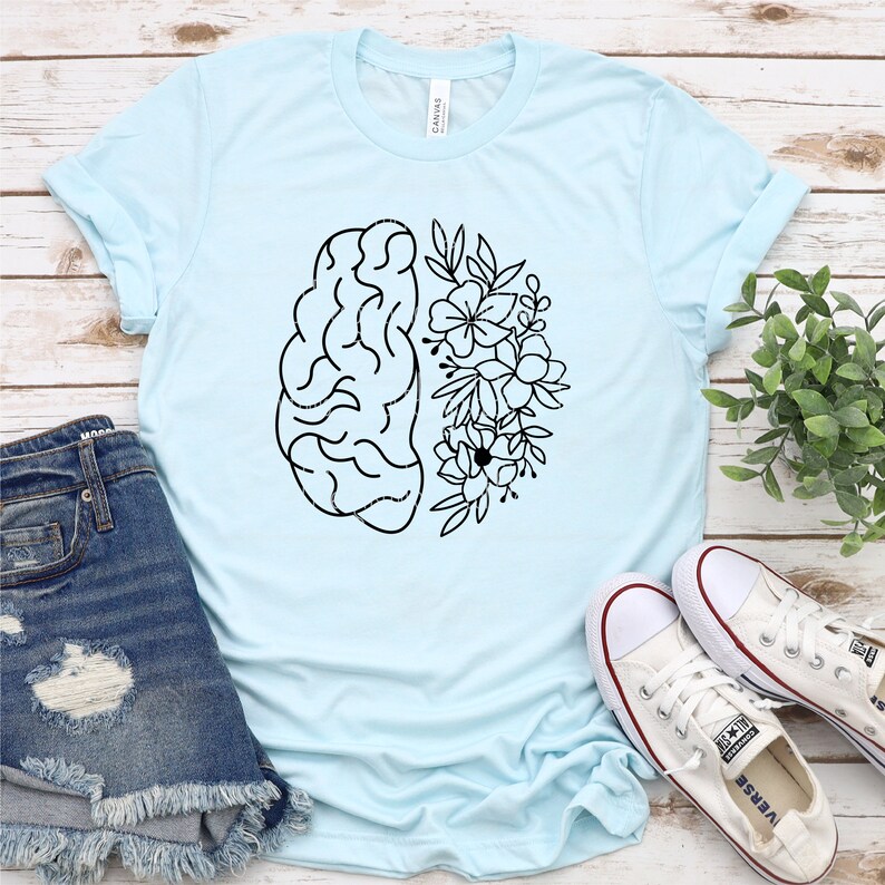 May include: A light blue t-shirt with a black and white graphic of a brain with flowers on one side. The shirt is laid flat on a wooden surface with a pair of white sneakers and a pair of blue denim shorts.