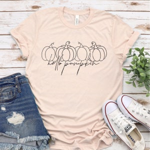 Pode incluir: Uma camiseta rosa claro com um contorno preto de ab&oacute;boras e o texto "hello pumpkin".