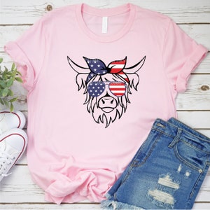 Puede incluir: Una camiseta rosa con una ilustraci&oacute;n en blanco y negro de una vaca de las tierras altas con gafas de sol y una bandana roja, blanca y azul. Las gafas de sol y la bandana est&aacute;n decoradas con la bandera estadounidense.
