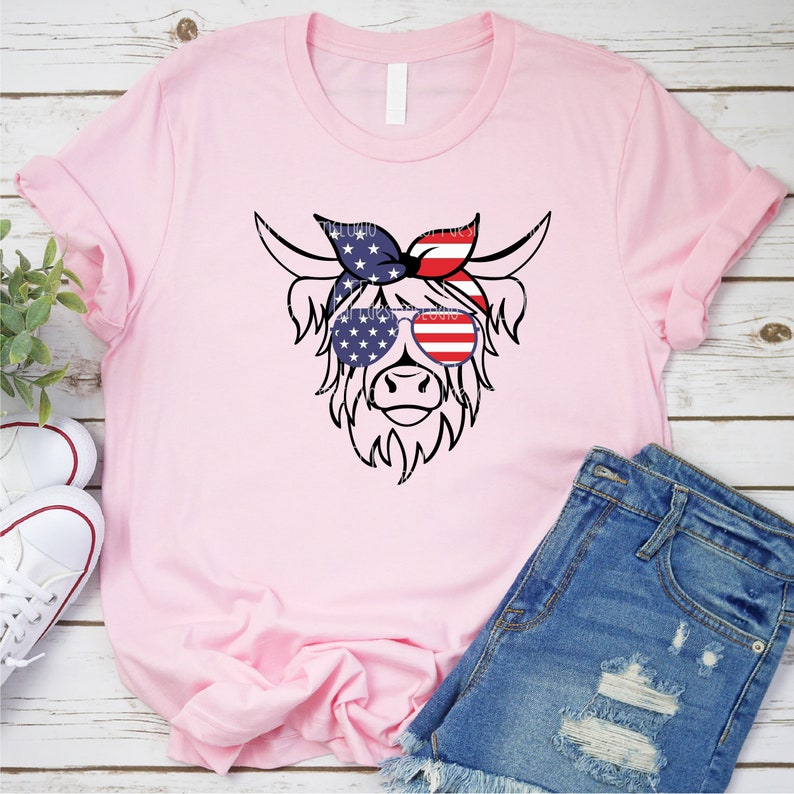 Puede incluir: Una camiseta rosa con una ilustraci&oacute;n en blanco y negro de una vaca de las tierras altas con gafas de sol y una bandana roja, blanca y azul. Las gafas de sol y la bandana est&aacute;n decoradas con la bandera estadounidense.