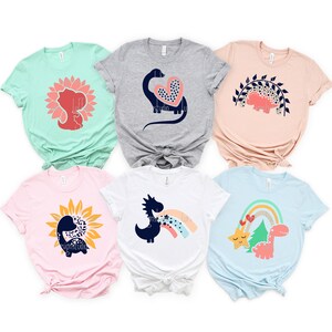 May include: Six different t-shirts featuring various dinosaur designs. The designs include a dinosaur with a heart, a dinosaur in a sunflower, a dinosaur with a rainbow, and a dinosaur with a star. The shirts are in various colors, including pink, blue, green, and gray.