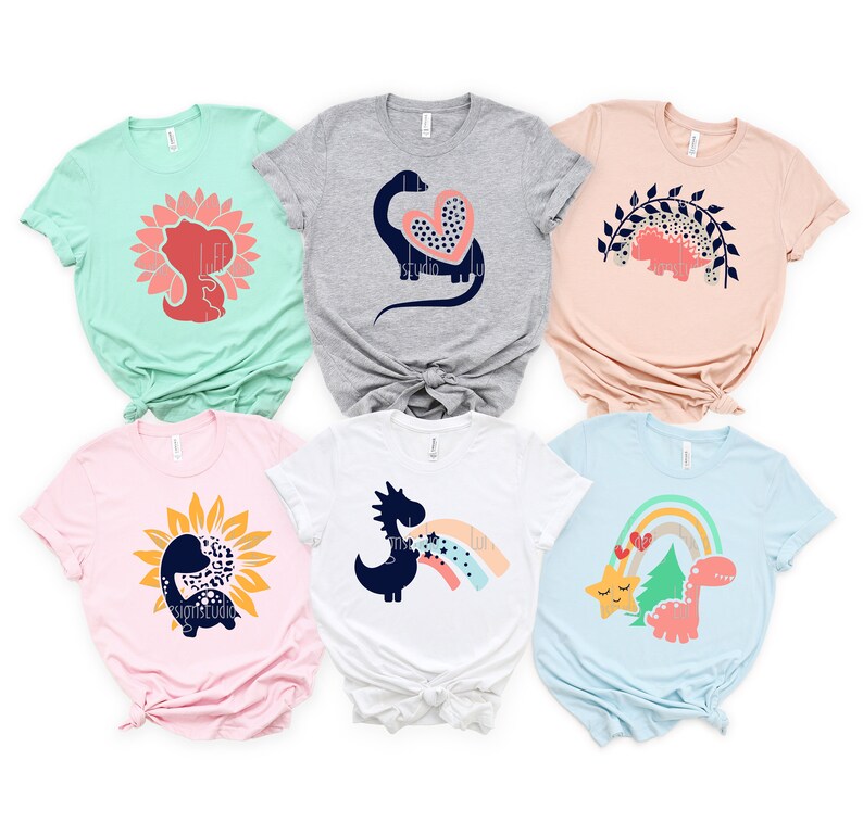 May include: Six different t-shirts featuring various dinosaur designs. The designs include a dinosaur with a heart, a dinosaur in a sunflower, a dinosaur with a rainbow, and a dinosaur with a star. The shirts are in various colors, including pink, blue, green, and gray.