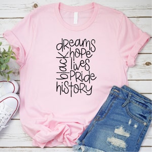 May include: Pink t-shirt with black text that reads "dreams hope black lives pride history".