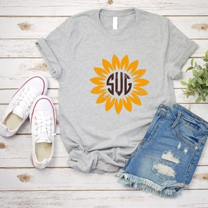 Puede incluir: Una camiseta gris con un monograma marr&oacute;n "SVG" dentro de un gr&aacute;fico de girasol amarillo. La camiseta est&aacute; colocada sobre un fondo de madera blanca con un par de zapatillas blancas y pantalones cortos de mezclilla azul.