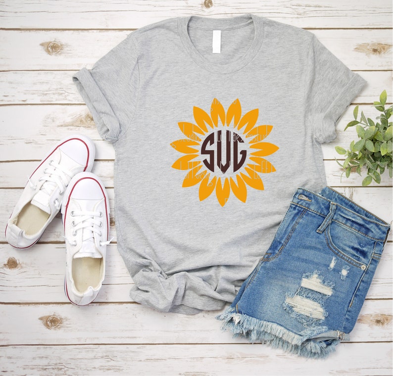 Puede incluir: Una camiseta gris con un monograma marr&oacute;n "SVG" dentro de un gr&aacute;fico de girasol amarillo. La camiseta est&aacute; colocada sobre un fondo de madera blanca con un par de zapatillas blancas y pantalones cortos de mezclilla azul.
