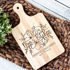 Puede incluir: Una tabla de cortar de madera con un dise&ntilde;o floral y el texto "Mom's Kitchen Breakfast, Lunch, Dinner open 24 hours".