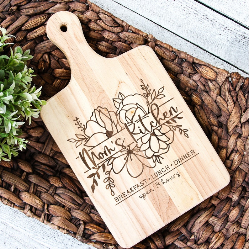 Puede incluir: Una tabla de cortar de madera con un dise&ntilde;o floral y el texto "Mom's Kitchen Breakfast, Lunch, Dinner open 24 hours".