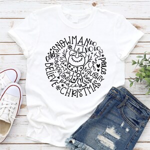 Op de afbeelding: Wit T-shirt met een zwart-wit ontwerp van een sneeuwpop omringd door woorden zoals "snowman", "ho ho ho", "peace", "believe", "joy", "kind", "naughty", en "christmas".