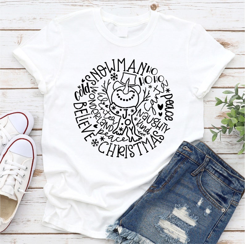 Op de afbeelding: Wit T-shirt met een zwart-wit ontwerp van een sneeuwpop omringd door woorden zoals "snowman", "ho ho ho", "peace", "believe", "joy", "kind", "naughty", en "christmas".