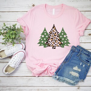 K&ouml;nnte beinhalten: Ein hellrosa T-Shirt mit einem Weihnachtsbaum-Design. Der zentrale Baum hat ein Leopardenmuster, flankiert von zwei gr&uuml;nen B&auml;umen mit wei&szlig;en Punkten. Das Shirt wird mit Jeansshorts und wei&szlig;en Turnschuhen gezeigt.
