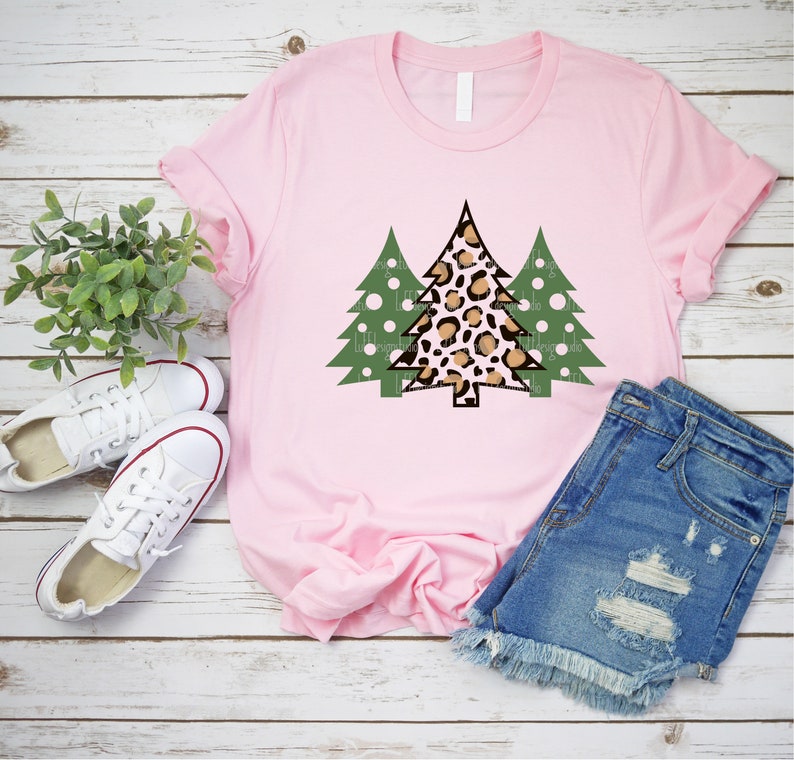 K&ouml;nnte beinhalten: Ein hellrosa T-Shirt mit einem Weihnachtsbaum-Design. Der zentrale Baum hat ein Leopardenmuster, flankiert von zwei gr&uuml;nen B&auml;umen mit wei&szlig;en Punkten. Das Shirt wird mit Jeansshorts und wei&szlig;en Turnschuhen gezeigt.