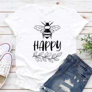 May include: White t-shirt with a black bee graphic and the word "HAPPY" printed below the bee. A leafy branch design is below the word "HAPPY".