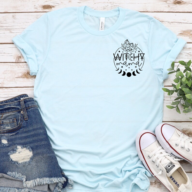 May include: A light blue t-shirt with a black graphic design of a circle with the text "Witchy mama" inside. The graphic also includes a crescent moon, stars, and crystals.
