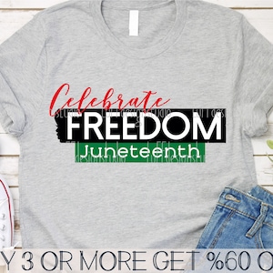 Puede incluir: Una camiseta gris con el texto "Celebrate FREEDOM Juneteenth" en rojo, negro y verde. El texto está en una fuente en negrita y de bloque. La camiseta está sobre una superficie de madera.