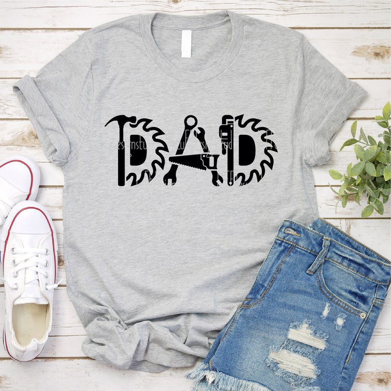 May include: A grey t-shirt with the word "DAD" spelled out using tools such as a hammer, saw, wrench, and a saw blade. The shirt is laid out on a woodgrain surface with a pair of white trainers and denim shorts.