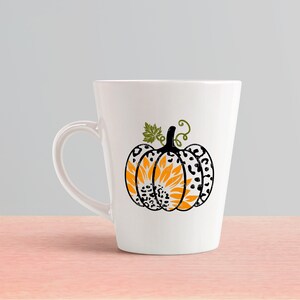 K&ouml;nnte beinhalten: Wei&szlig;e Keramiktasse mit einem schwarzen und wei&szlig;en Leopardenmuster-K&uuml;rbis mit einer Sonnenblume im Inneren. Der K&uuml;rbis ist orange und gelb.