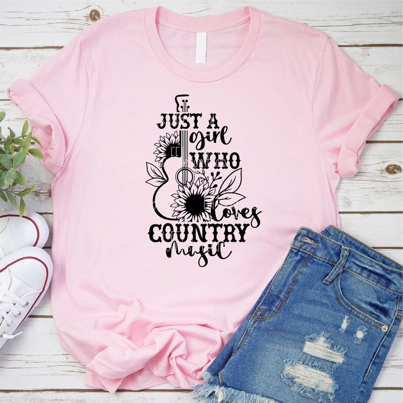 Puede incluir: Camiseta rosa claro con mangas enrolladas que presenta un gr&aacute;fico negro de una guitarra, girasoles y el texto "Just a girl who loves country music". La camiseta se muestra con pantalones cortos de mezclilla y zapatillas blancas.