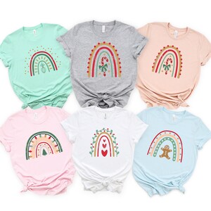 May include: Six different Christmas-themed rainbow t-shirts in various colors. Each shirt features a different design, including a candy cane, a Christmas tree, a gingerbread man, and hearts.