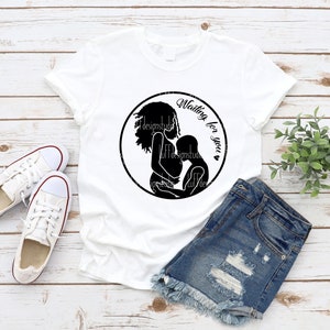 May include: White t-shirt with a black silhouette of a pregnant woman holding a child. The text "Waiting for you" is written in a circle around the silhouette.