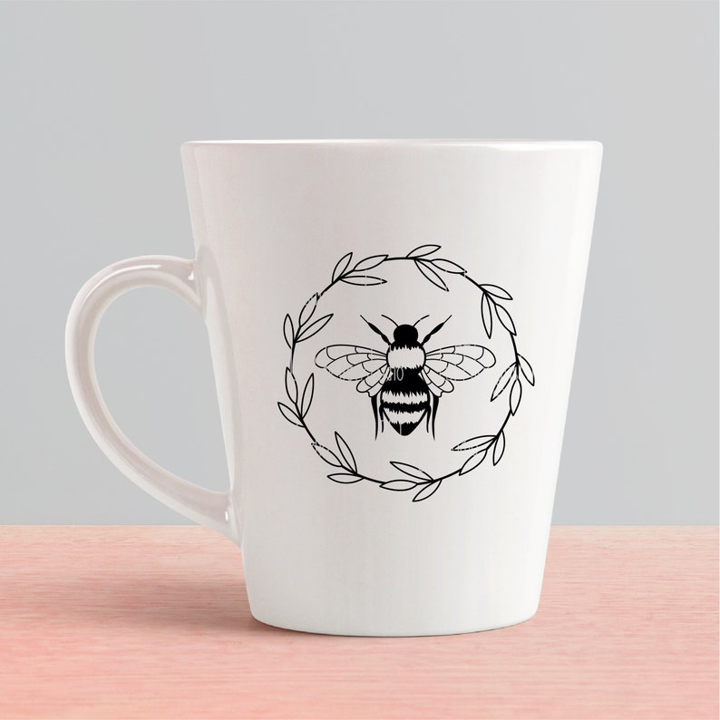 May include: White ceramic mug featuring a black and white illustration of a bee within a wreath of leaves.