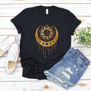 May include: A black t-shirt with a gold glitter design of a sun and moon with stars and a crescent moon. The design is on a black t-shirt with a white background.
