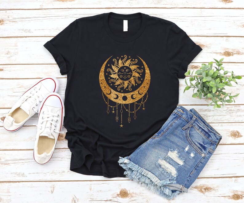 May include: A black t-shirt with a gold glitter design of a sun and moon with stars and a crescent moon. The design is on a black t-shirt with a white background.
