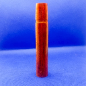 May include: A cylindrical wooden object with a smooth, polished finish and a rich reddish-brown grain pattern. The item features a slightly tapered top section and a straight, uniform body, showcasing the natural texture and warm tones of the wood.