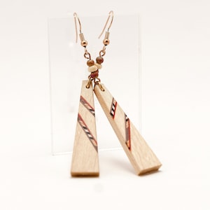 Puede incluir: Un par de pendientes de madera con diseño geométrico. Los pendientes son de color marrón claro con detalles rojos, negros y blancos. Cuelgan de ganchos de color cobre y están conectados por pequeñas cuentas. Los pendientes tienen forma triangular.