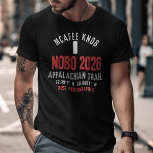 May include: Black crew neck t-shirt with white and red text. The text reads "MCAFEE KNOB NOBO 2026 APPALACHIAN TRAIL 37.3919° N, 80.0867° W MOST PHOTOGRAPHED". The shirt is worn by a person.