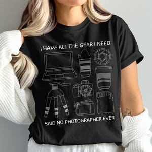 May include: A black short-sleeved t-shirt featuring white line art of photography equipment including a laptop, camera, lens, tripod, and memory card. The text reads I HAVE ALL THE GEAR I NEED at the top and SAID NO PHOTOGRAPHER EVER at the bottom.