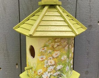 Peut inclure: Un nichoir en bois vert avec un toit en bardeaux et un imprimé de marguerites sur le devant. Le nichoir a une ouverture ronde pour que les oiseaux puissent entrer.