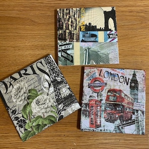 May include: Three decorative square tiles with a vintage aesthetic. The tiles feature images of Paris, New York City, and London. The Paris tile has a black and white image of the Eiffel Tower with white roses and the text "Paris 1900". The New York City tile has a collage of images including the Brooklyn Bridge, a street sign that says "Time Square", and a street sign that says "One Way". The London tile has a red double-decker bus, a red telephone booth, and the text "London" and "Underground".