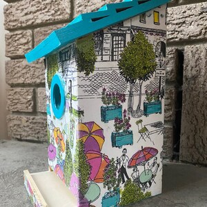 May include: A turquoise blue birdhouse with a white base and a roof with a scalloped edge. The birdhouse is decorated with a black and white illustration of a Parisian street scene with umbrellas, trees, and buildings. The text "PARIS" is visible on the side of the birdhouse.