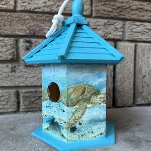 May include: A turquoise wooden birdhouse featuring a turtle design. The birdhouse has a pitched roof, a circular entry hole, and a perch. A white rope is attached to the top for hanging. The birdhouse is painted with a sea turtle and ocean scene.