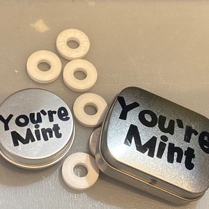 May include: A silver tin with the text "You're Mint" on it.  There are several white round mints next to the tin.