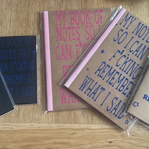 Può includere: Cinque piccoli quaderni con frasi divertenti stampate sulla copertina. I quaderni sono avvolti in plastica trasparente e hanno copertine marroni e nere. Le frasi includono "My Book of Notes So I Can Remember What I Said", "My Book of Notes So I Can F'cking Remember What I Said", "My Notebook So I Can F'cking Remember What I Said", e "My Book of Passwords Can Never F'cking Remember".